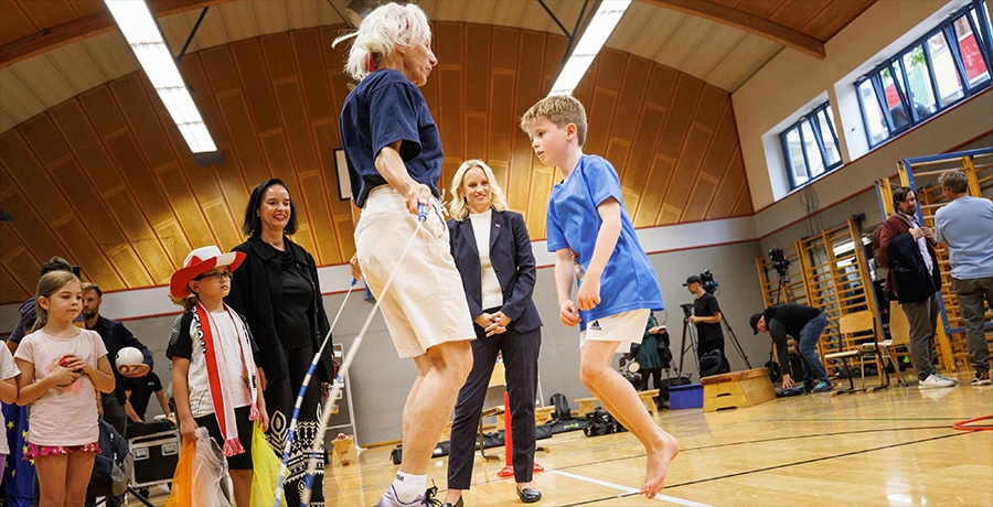 Ältere Frau und Kind beim Springschnurspiel; im Hintergrund eine Turnhalle und StS Schmidt