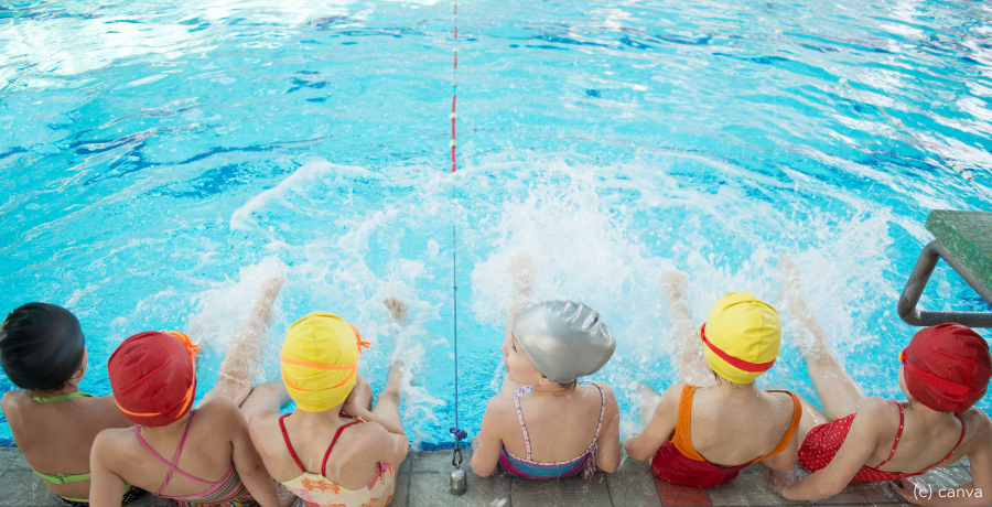 Sechs Kinder mit Badehaube sitzen am Rand von Schwimmbecken 