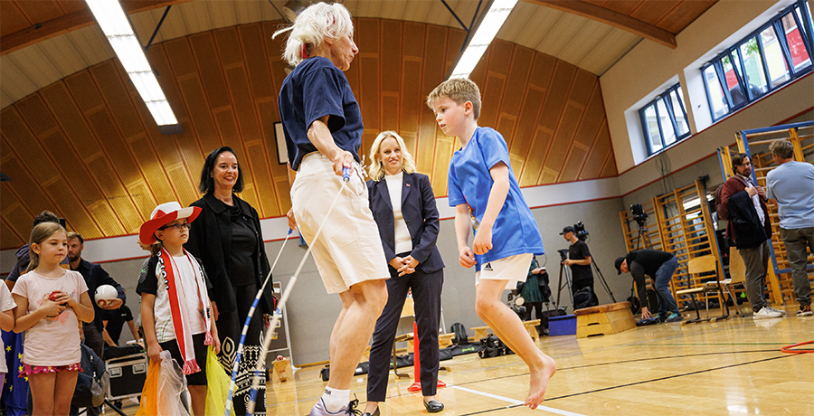 Ältere Frau und Kind beim Springschnurspiel; im Hintergrund eine Turnhalle und StS Schmidt