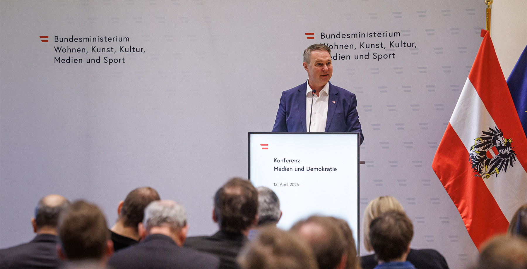 Andreas Babler auf Podium bei Konferenz "Medien und Demokratie" am 13. April 2026. Österreich-Flagge neben Bühne.